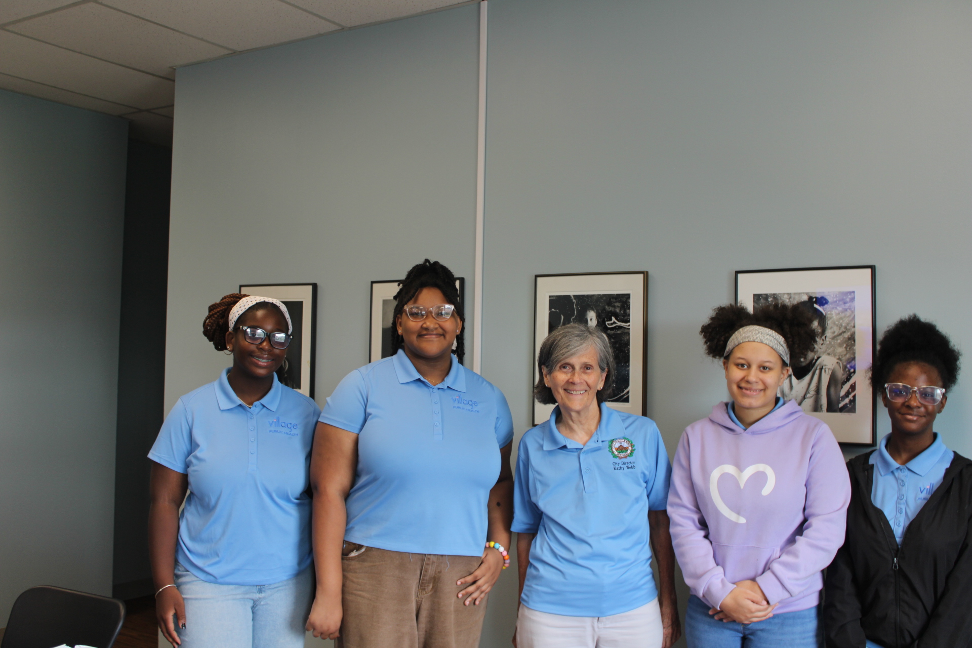 Four young Black women from the Village Public Health Summer Institute cohort standing shoulder-to-shoulder in matching light blue polos with Little Rock City Director Kathy Webb inside Little Rock City Hall during a civic engagement visit.