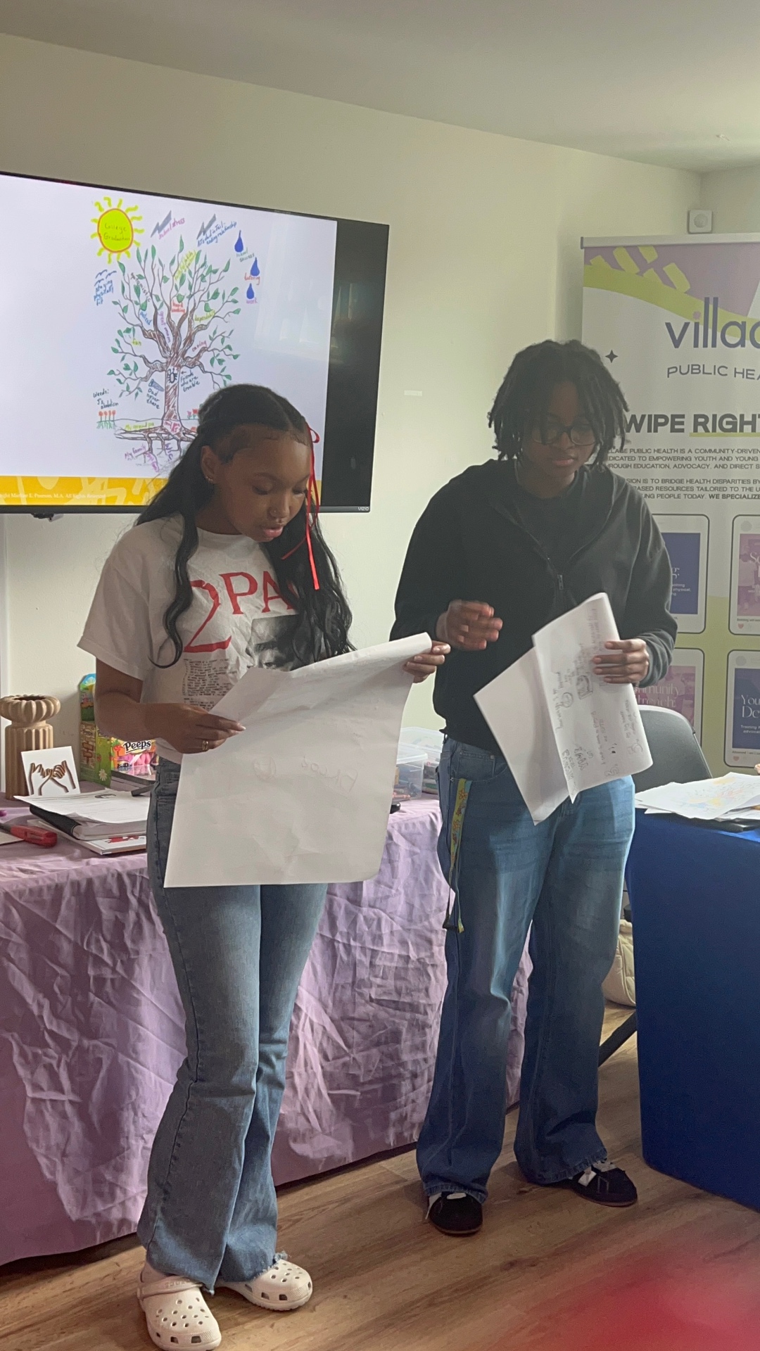 Two young Black women in the 2026 Village Public Health Summer Institute cohort standing in front of a classroom monitor in Little Rock, presenting large hand-drawn Tree of Life posters that map their roots, stressors, and hopes.